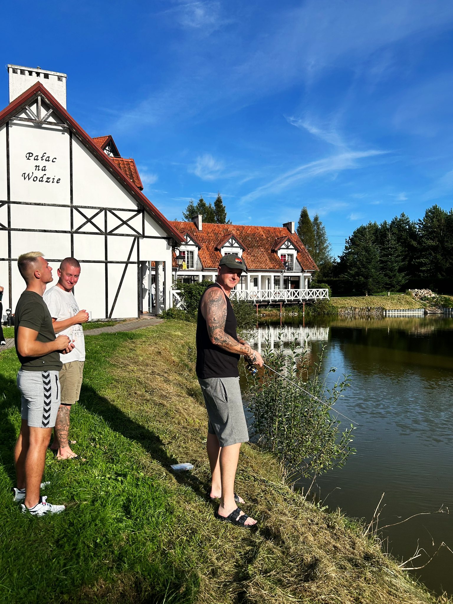 Three men standing by a pond near a building labeled "Pałac na Wodzie."