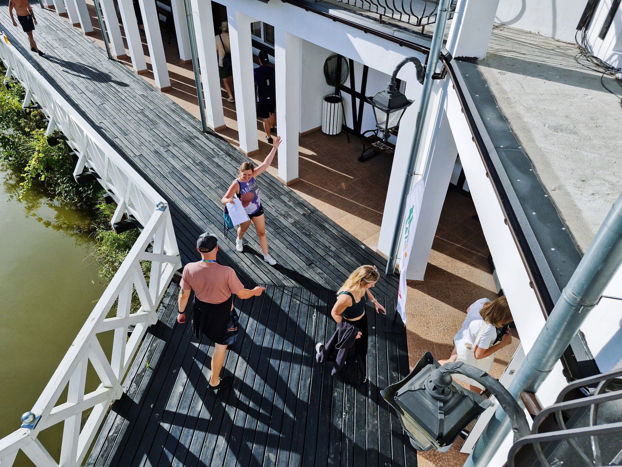 People walking on a wooden boardwalk beside a white building on a sunny day.