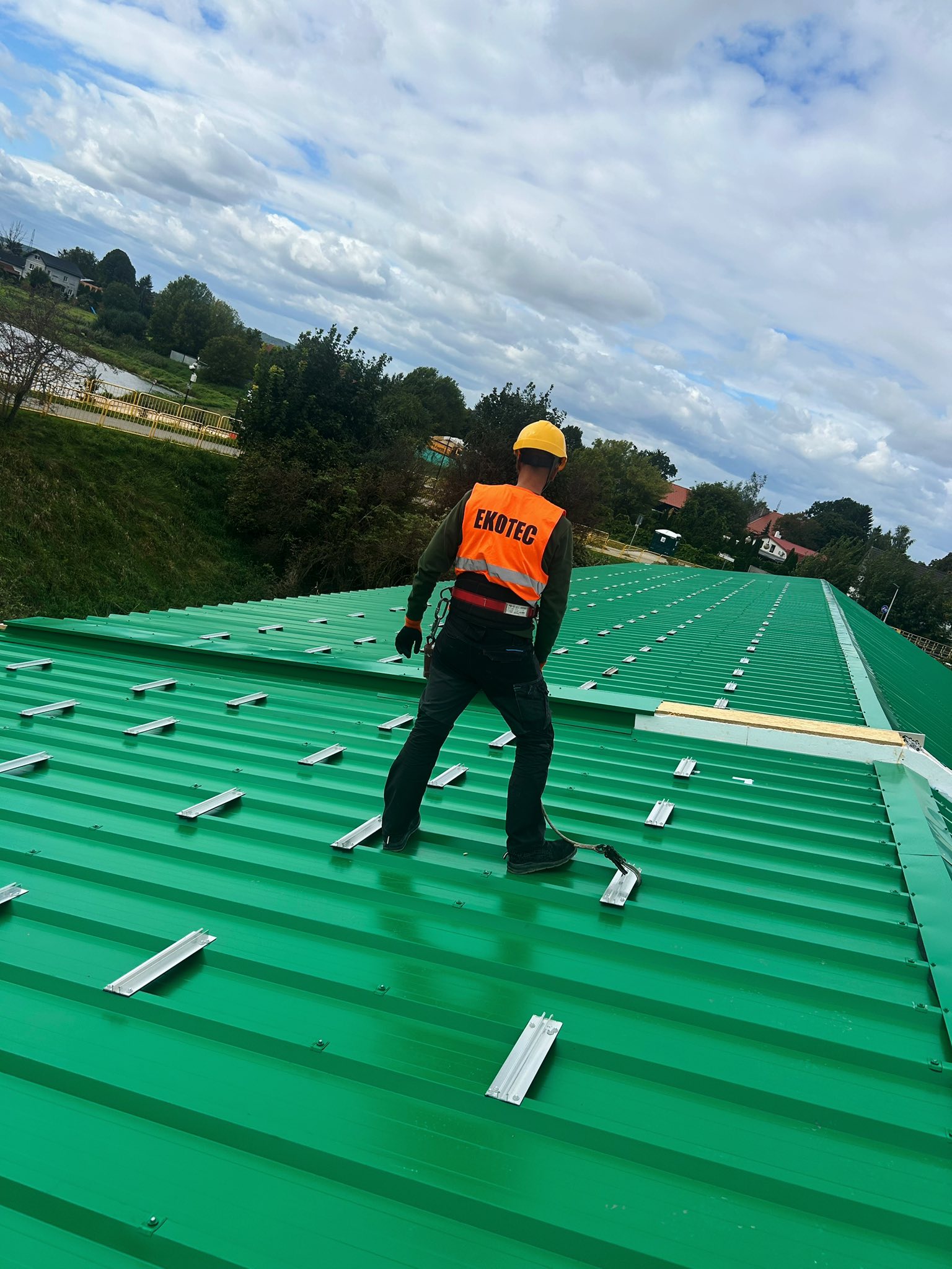 Worker in orange vest and helmet on green metal roof under cloudy sky.