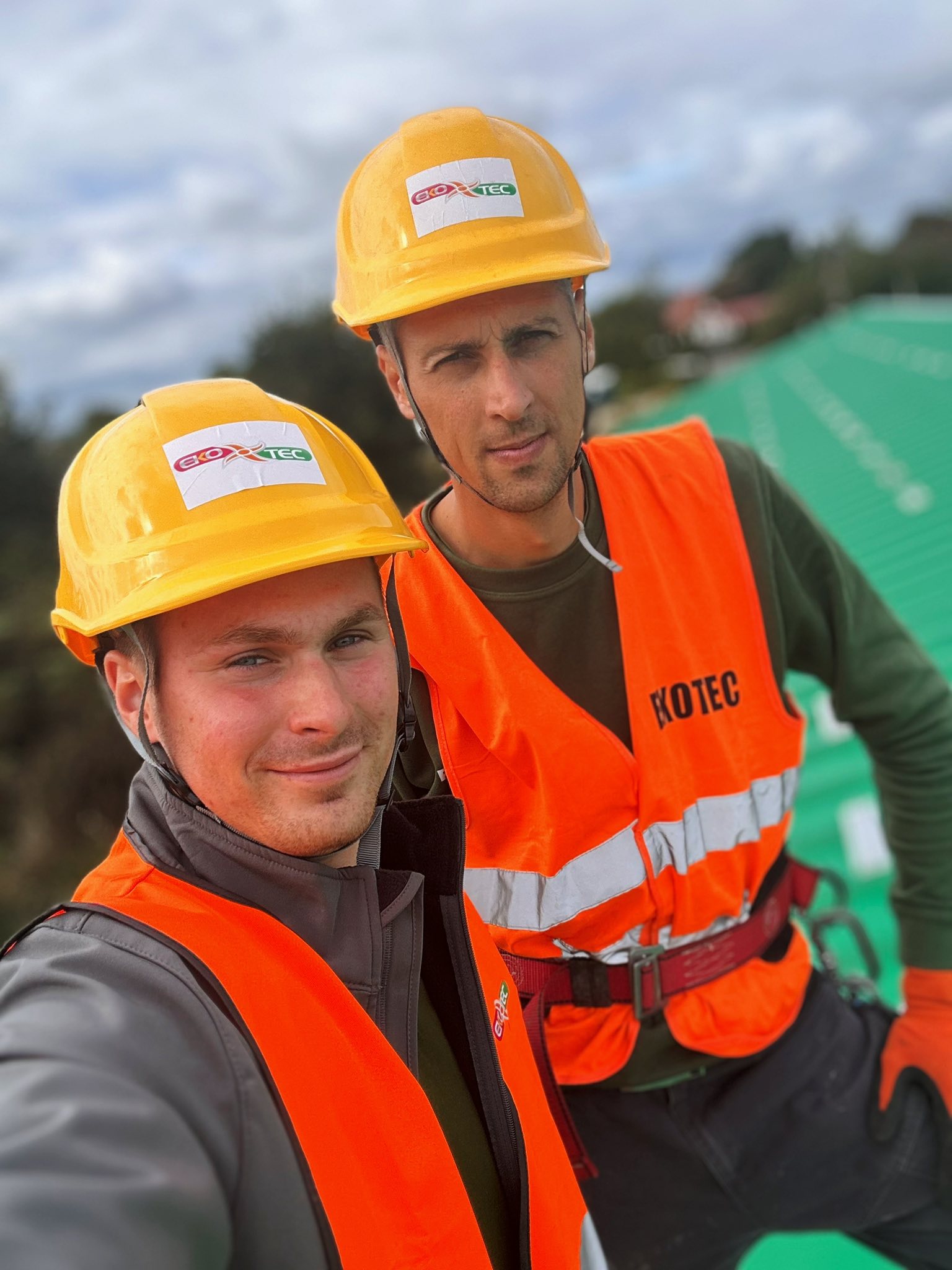 Two construction workers in orange vests and yellow helmets on a rooftop.