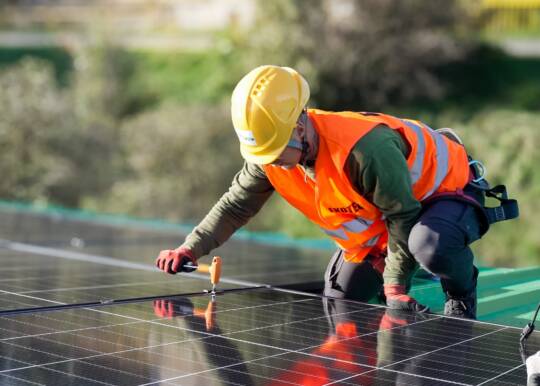 Worker in safety gear installing solar panels on a rooftop in sunlight.