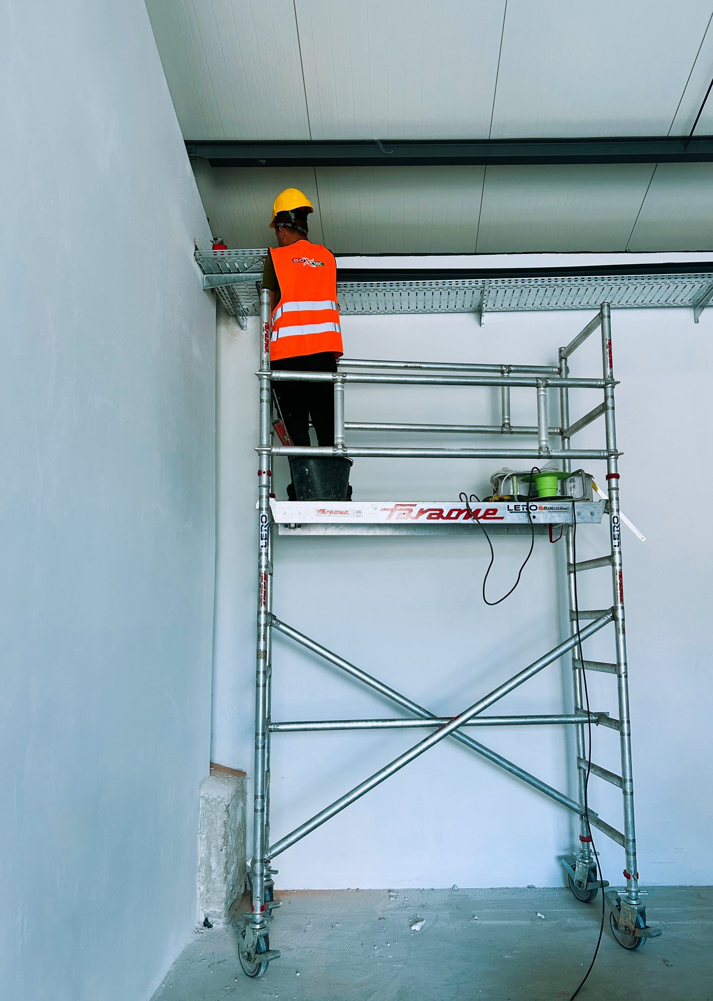 Construction worker on scaffolding installing ceiling panels in a large room.