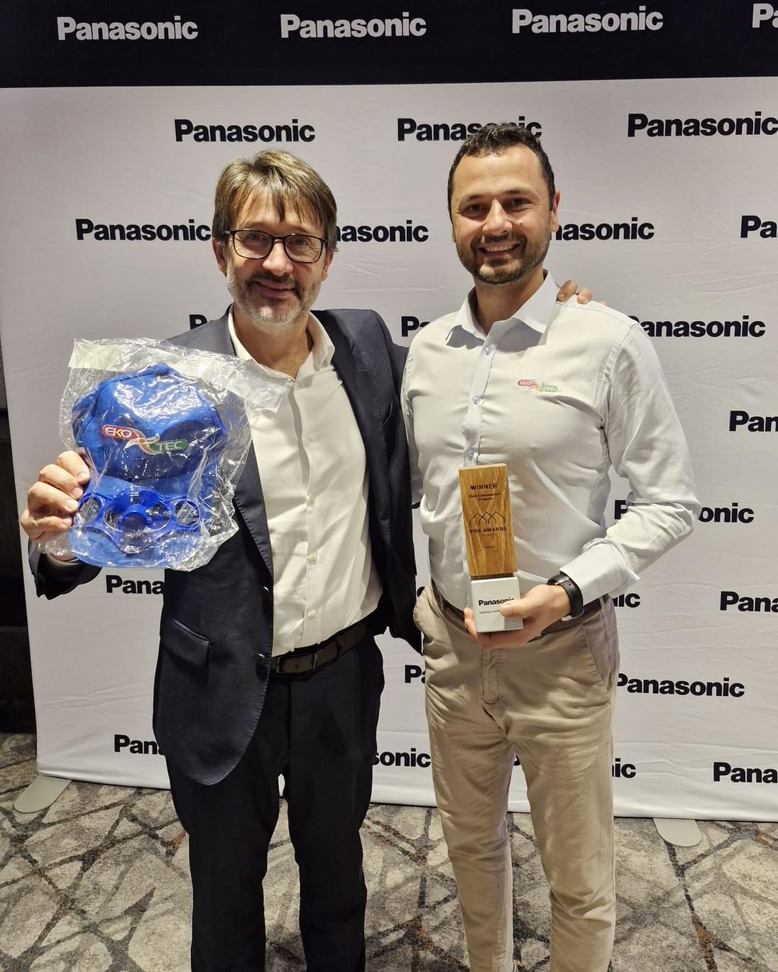 Two men smiling, holding awards, in front of Panasonic-branded backdrop.