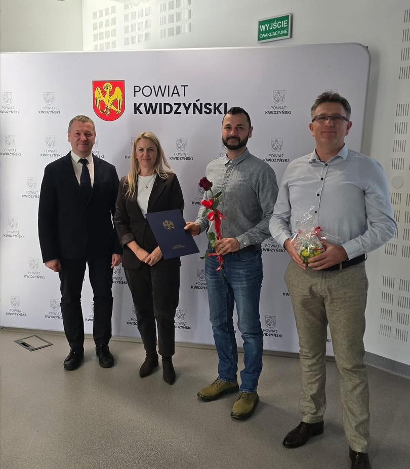 Four people standing in front of a "Powiat Kwidzyński" backdrop, holding certificates and gifts.