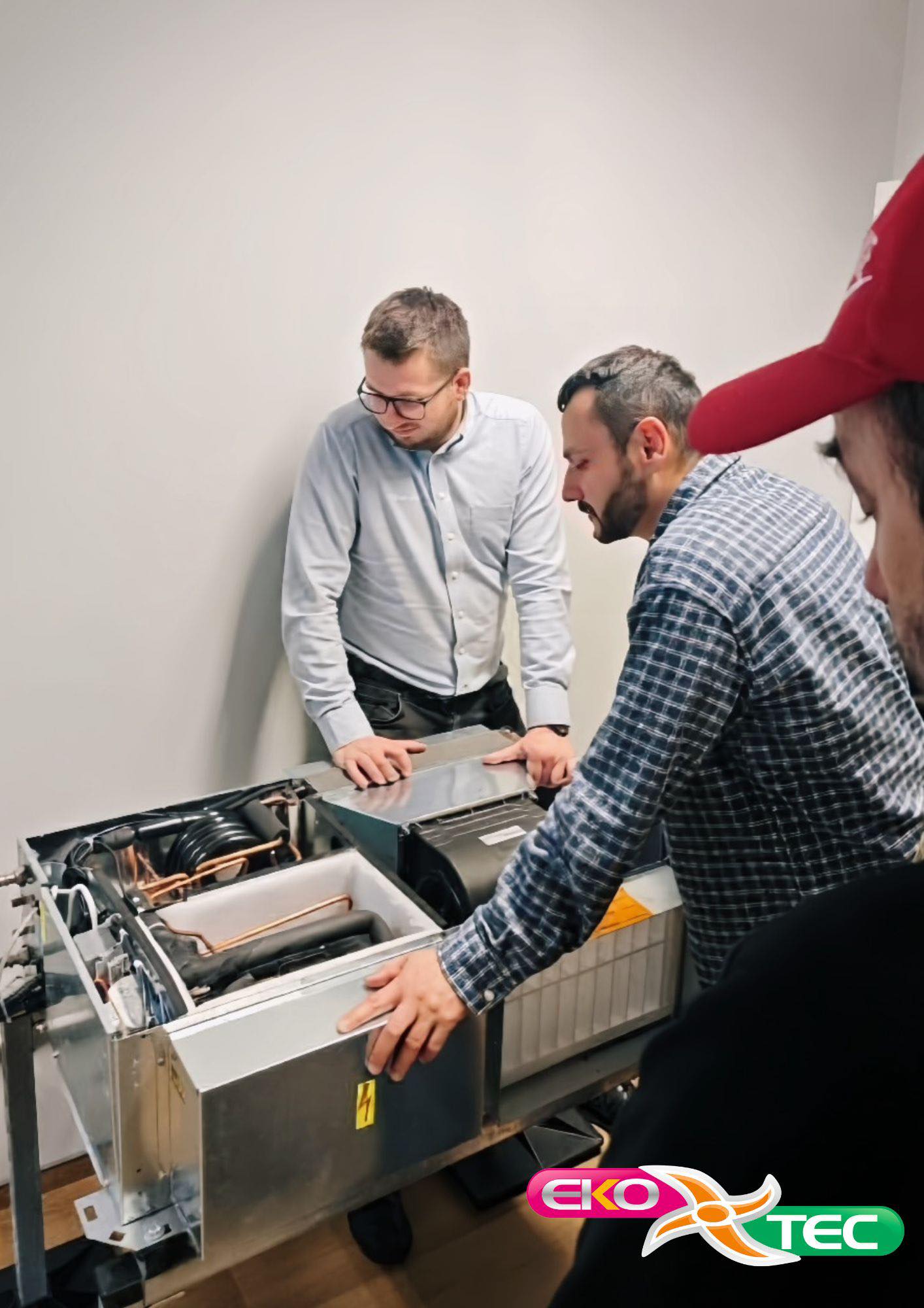 Three men examining HVAC equipment in a training room, Eko-Tec logo visible.