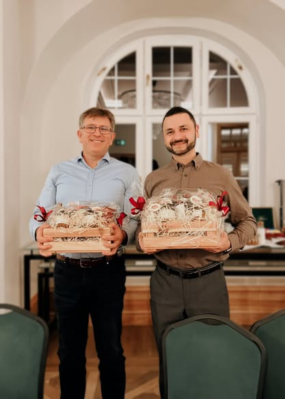 Two men smiling, holding gift baskets with red bows in a decorated room.