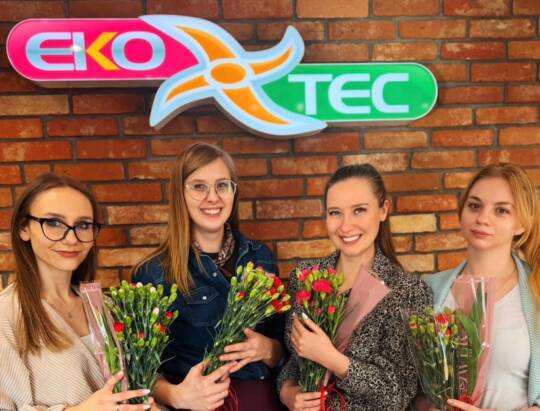 Four women holding bouquets, smiling in front of EKOTEC sign on brick wall.