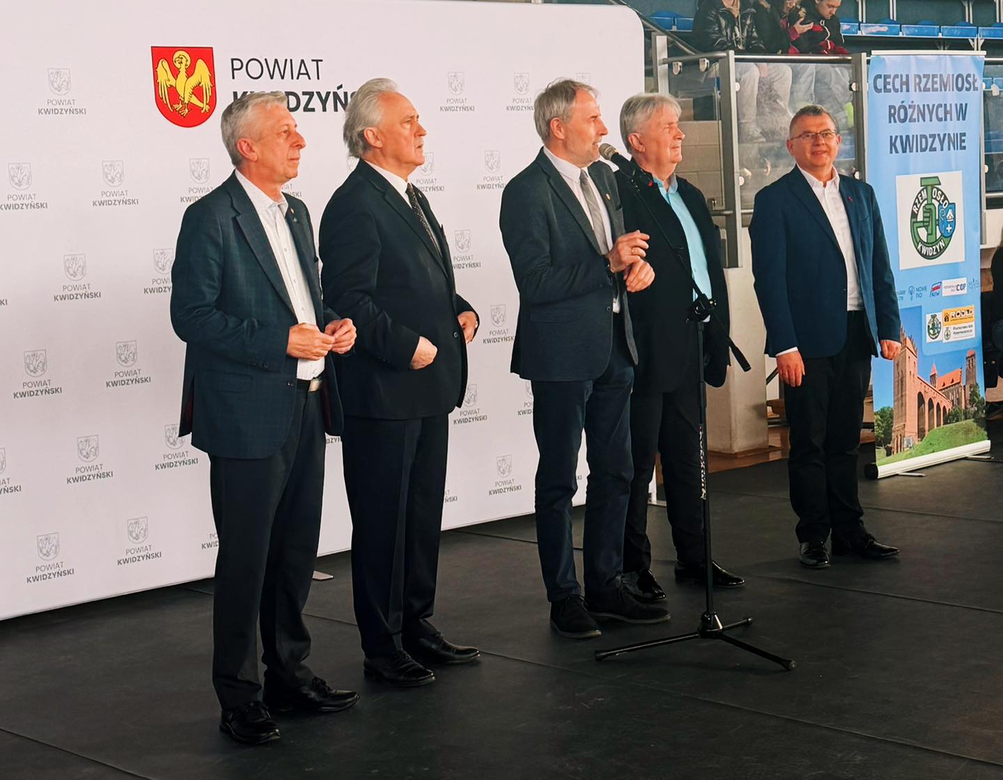 Five men in suits standing on stage at Kwidzyn educational fair.