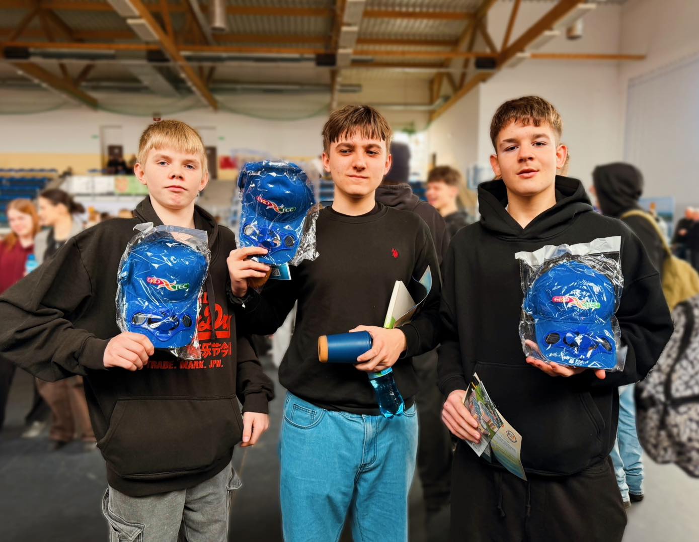 Three young people holding blue caps in a busy indoor setting.