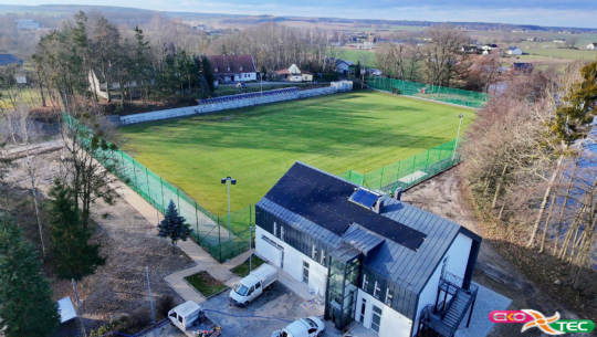 Aerial view of a fenced soccer field with adjacent modern building and parked cars.