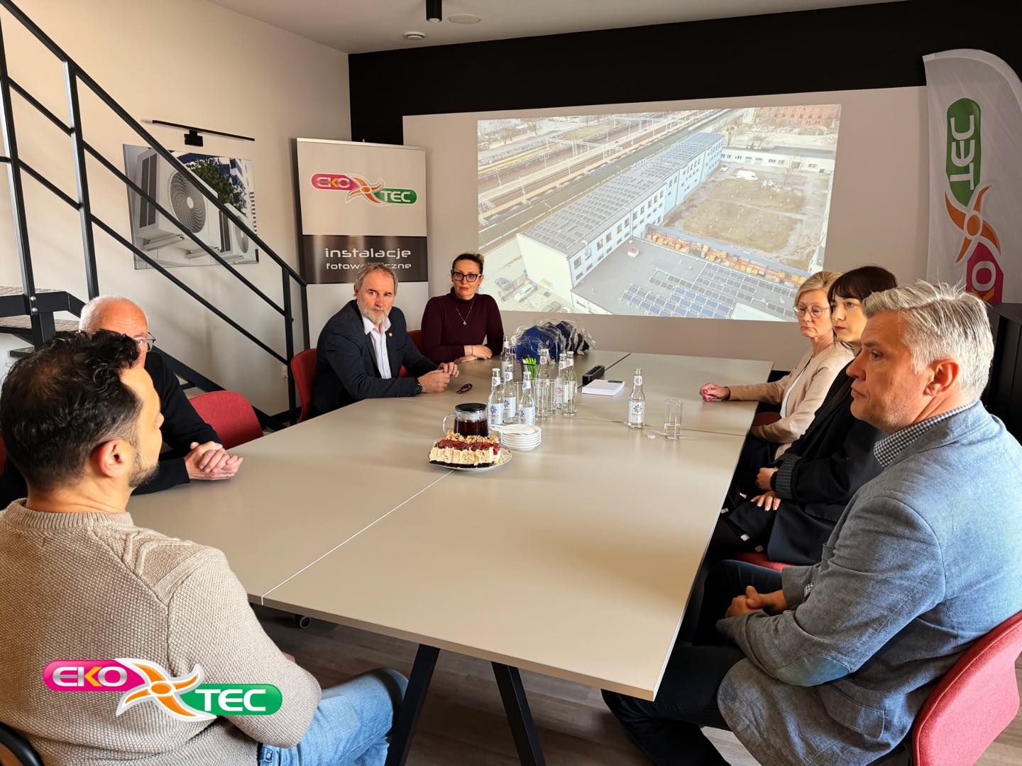 Seven people in a meeting room with a presentation screen and logo banners.