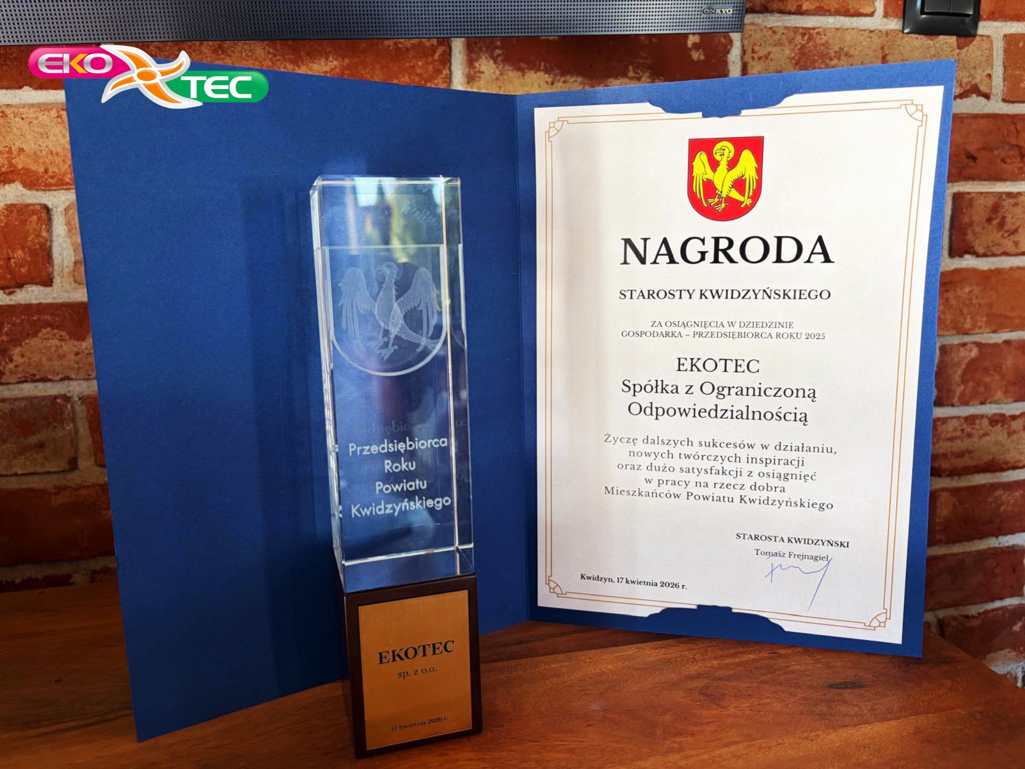 Award plaque and certificate for EKOTEC on a wooden table with brick background.